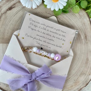 May include: A light purple ribbon tied in a bow adorns an open envelope. Inside, a keychain with the word "TATA" is visible, along with a card with French text. The background features daisies and greenery.