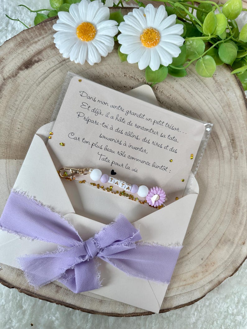 May include: A light purple ribbon tied in a bow adorns an open envelope. Inside, a keychain with the word "TATA" is visible, along with a card with French text. The background features daisies and greenery.