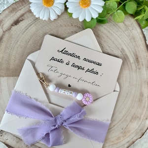 May include: A light beige envelope with a lavender ribbon and a card inside. The card has text in French. A beaded charm with the letters "TATA" and a small purple flower is attached to the envelope.