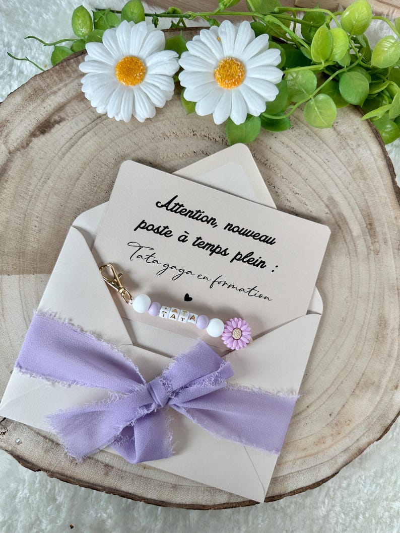 May include: A light beige envelope with a lavender ribbon and a card inside. The card has text in French. A beaded charm with the letters "TATA" and a small purple flower is attached to the envelope.