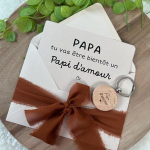 May include: A greeting card in an envelope with a brown ribbon, and a wooden keychain. The card reads "PAPA tu vas &ecirc;tre bient&ocirc;t un Papi d'amour". The keychain is round and has the words "Papi d'amour" on it.