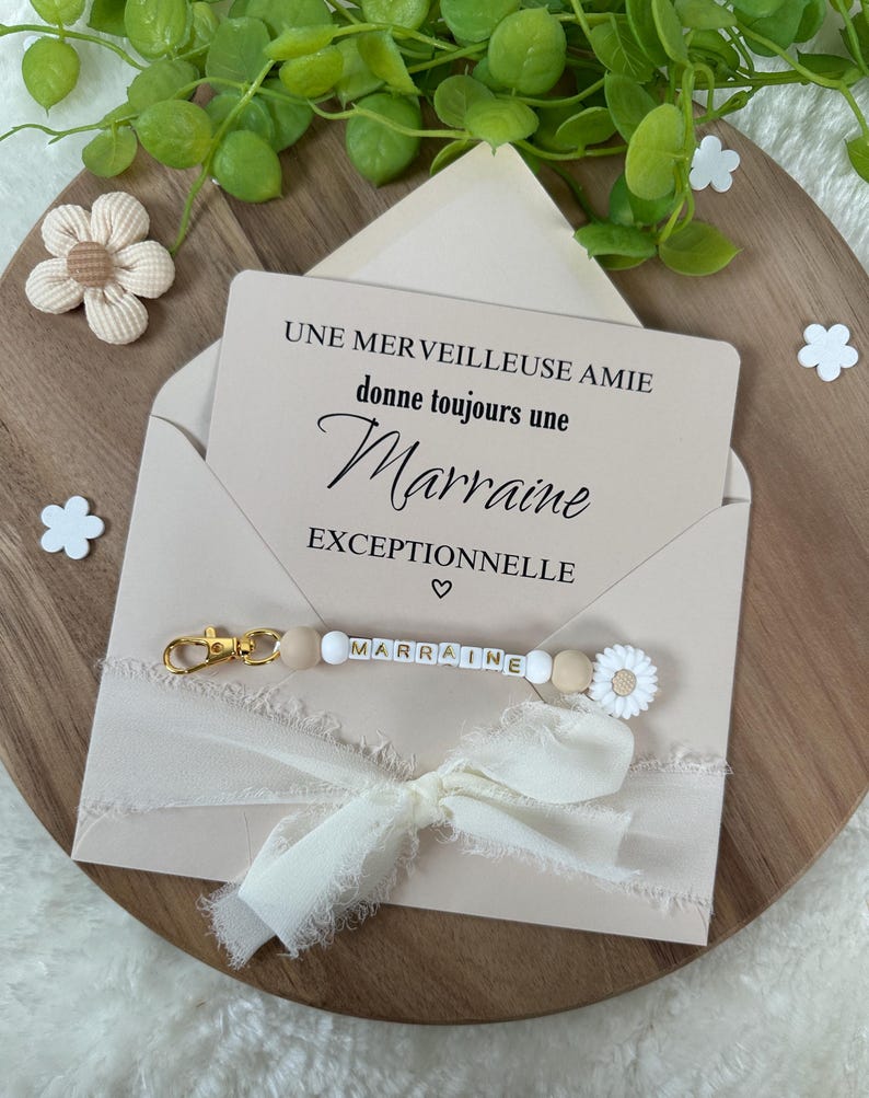 May include: A pink envelope with a white card inside that reads "Une merveilleuse amie donne toujours une Marraine exceptionnelle". A white ribbon with a gold keychain with white beads and the word "Marraine" is attached to the envelope.