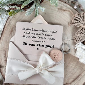 May include: A light pink card and envelope set with a cream-colored ribbon. The card reads "You're going to be a grandpa" in French. A wooden keychain with the words "Daddy of love" is also included.