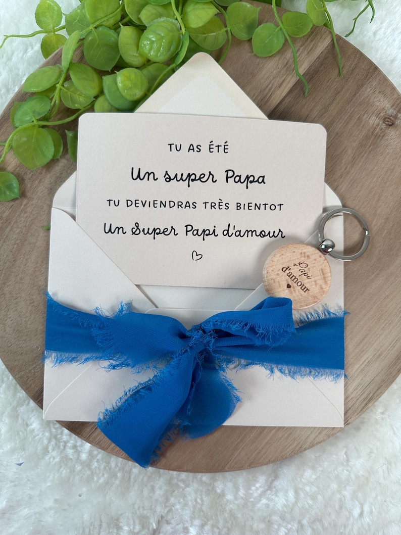 May include: A greeting card in an envelope with a blue ribbon. The card reads "TU AS &Eacute;T&Eacute; Un super Papa TU DEVIENDRAS TR&Egrave;S BIENTOT Un Super Papi d'amour". A wooden keychain with the words "Papi d'amour" is also included.