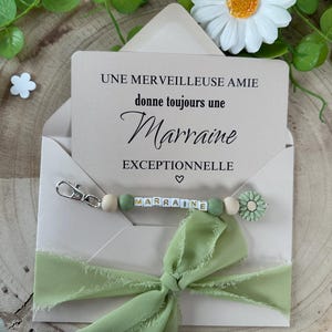 May include: A light brown envelope with a green ribbon bow and a keychain with green and white beads that spell "marraine". The envelope has a card inside that reads "Une merveilleuse amie donne toujours une marraine exceptionnelle".
