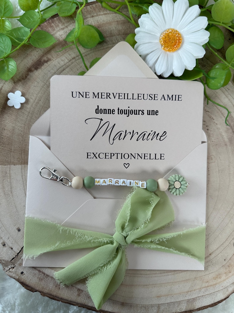 May include: A light brown envelope with a green ribbon bow and a keychain with green and white beads that spell "marraine". The envelope has a card inside that reads "Une merveilleuse amie donne toujours une marraine exceptionnelle".