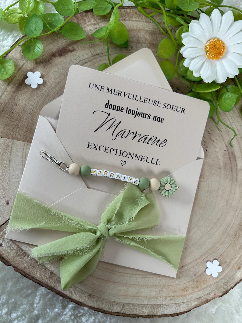 May include: A light green ribbon tied in a bow on top of a card that reads "Une merveilleuse soeur donne toujours une marraine exceptionnelle". The card is on a wooden surface with a green plant and white flowers.