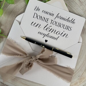May include: A cream-colored envelope with a beige ribbon bow, holding a card with French text and a black pen. The pen has gold accents and a small heart. The card reads "Un cousin formidable donne toujours un t&eacute;moin exceptionnel."