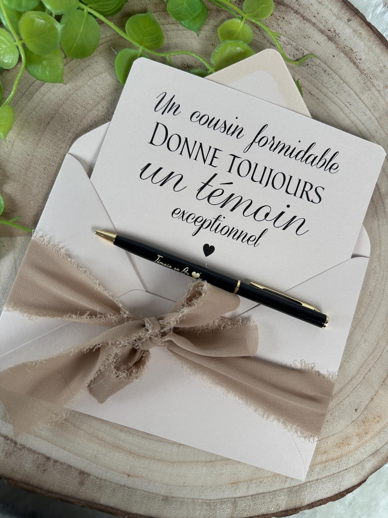 May include: A cream-colored envelope with a beige ribbon bow, holding a card with French text and a black pen. The pen has gold accents and a small heart. The card reads "Un cousin formidable donne toujours un t&eacute;moin exceptionnel."
