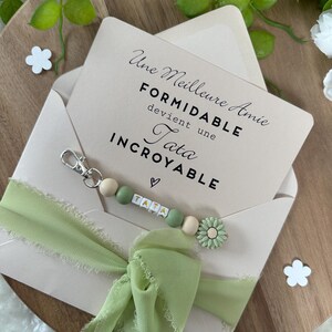 Puede incluir: Un sobre rosa claro con una cinta verde y un llavero de cuentas. El sobre tiene un mensaje que dice "Une Meilleure Amie Formidable devient une Tata Incroyable". El llavero tiene un dije de coraz&oacute;n y la palabra "Tata" escrita en cuentas.