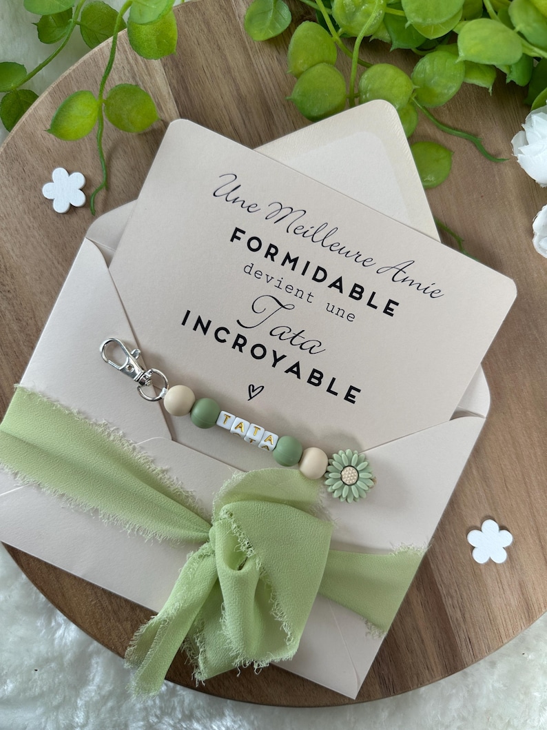 Puede incluir: Un sobre rosa claro con una cinta verde y un llavero de cuentas. El sobre tiene un mensaje que dice "Une Meilleure Amie Formidable devient une Tata Incroyable". El llavero tiene un dije de coraz&oacute;n y la palabra "Tata" escrita en cuentas.