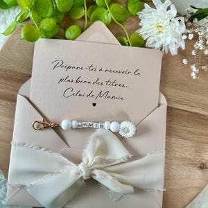 Pu&ograve; includere: Una busta beige con un nastro color crema e un portachiavi con la scritta "MAMIE" in perline di lettere. La busta contiene una carta con il testo "Pr&eacute;pare-toi &agrave; recevoir le plus beau des titres... Celui de Mamie."