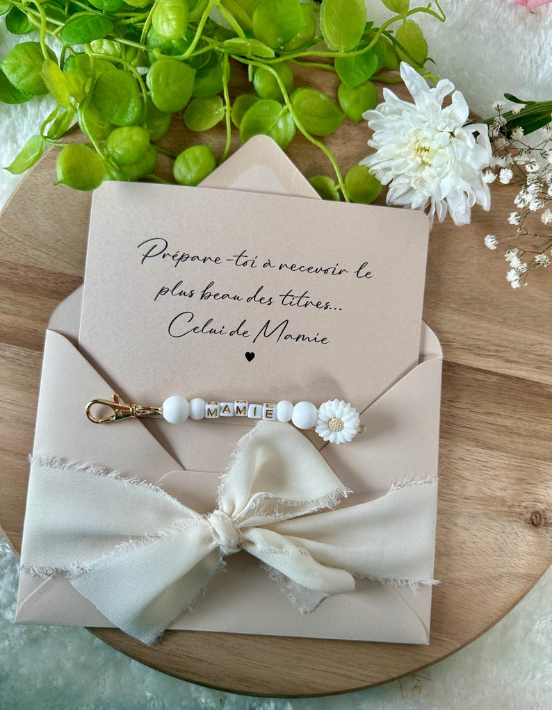 Pu&ograve; includere: Una busta beige con un nastro color crema e un portachiavi con la scritta "MAMIE" in perline di lettere. La busta contiene una carta con il testo "Pr&eacute;pare-toi &agrave; recevoir le plus beau des titres... Celui de Mamie."