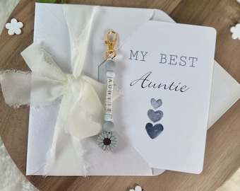 Può includere: Un biglietto bianco con il testo "My Best Auntie" e un design a cuore blu ad acquerello. Un portachiavi con perline bianche e grigie con la parola "Auntie" scritta in lettere dorate e un ciondolo a forma di fiore grigio è attaccato al biglietto.