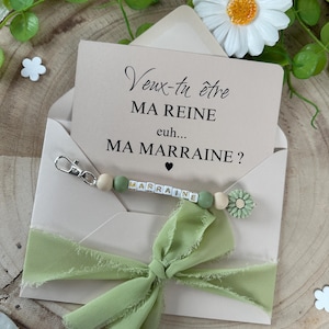 May include: A light green envelope with a white card inside. The card has the text "Veux-tu &ecirc;tre MA REINE euh... MA MARRAINE?" and a green and white beaded keychain with the word "MARRAINE" spelled out.