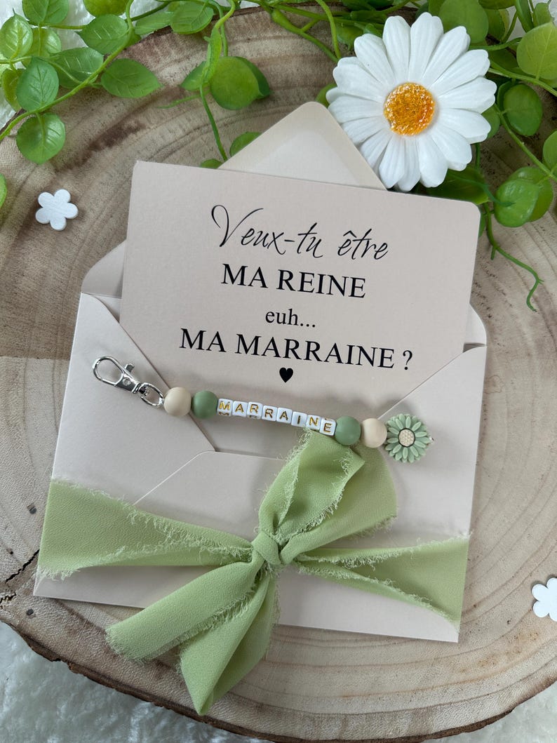 May include: A light green envelope with a white card inside. The card has the text "Veux-tu &ecirc;tre MA REINE euh... MA MARRAINE?" and a green and white beaded keychain with the word "MARRAINE" spelled out.