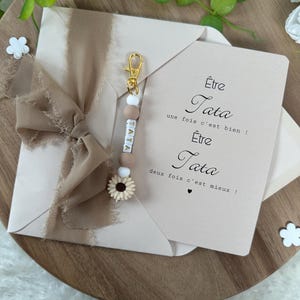 May include: A pink card with the text "&Ecirc;tre Tata une fois c'est bien! &Ecirc;tre Tata deux fois c'est mieux!"  A keychain with the word "TATA" spelled out in white beads with brown accents and a small sunflower charm.