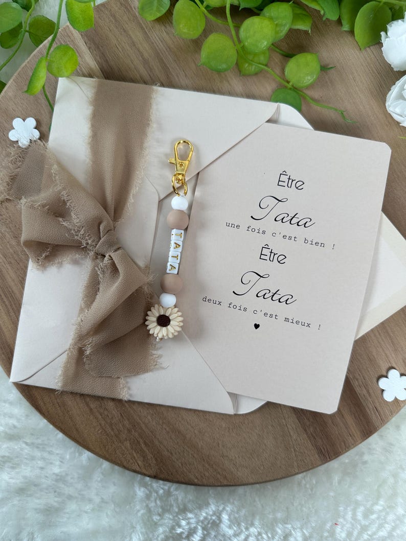 May include: A pink card with the text "&Ecirc;tre Tata une fois c'est bien! &Ecirc;tre Tata deux fois c'est mieux!"  A keychain with the word "TATA" spelled out in white beads with brown accents and a small sunflower charm.