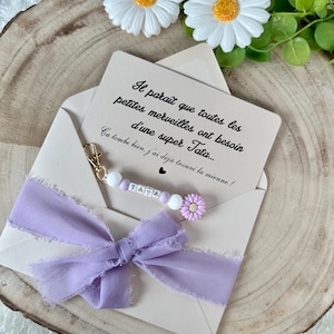 May include: A light beige envelope with a lavender ribbon bow and a beaded charm that spells "TATA". The envelope contains a card with French text and is decorated with white daisies with yellow centers.