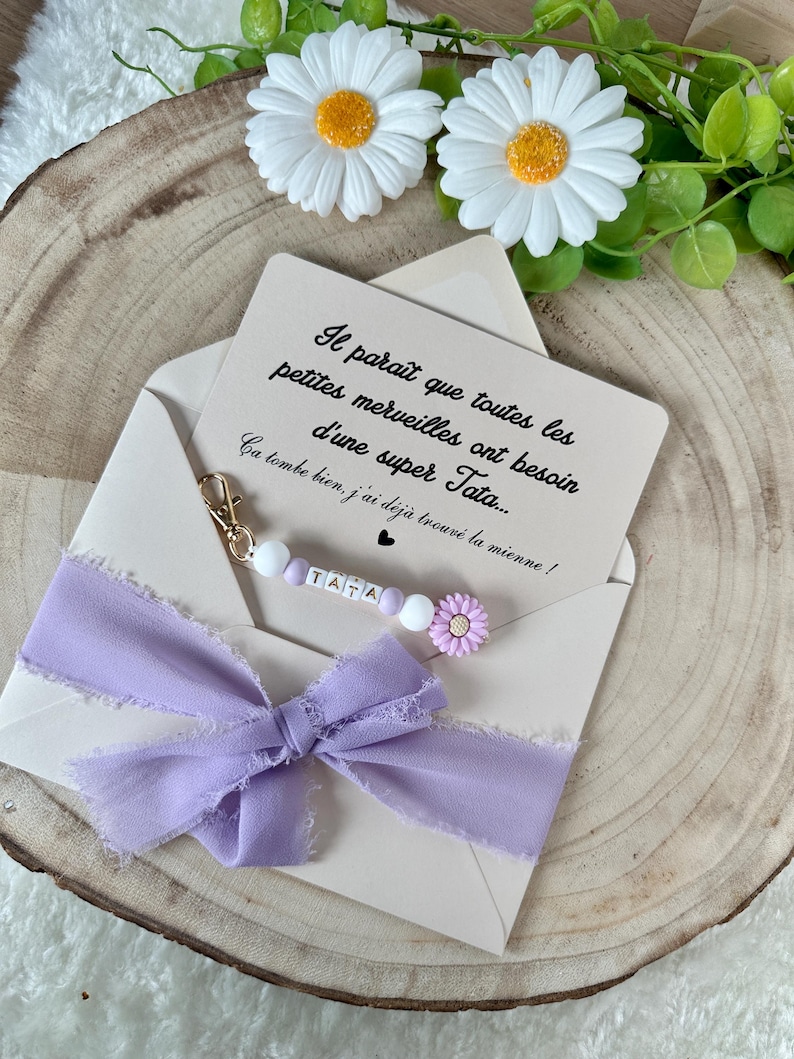 May include: A light beige envelope with a lavender ribbon bow and a beaded charm that spells "TATA". The envelope contains a card with French text and is decorated with white daisies with yellow centers.
