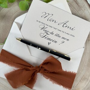 May include: An invitation card with the text "Mon Ami" and "Veux-tu &ecirc;tre mon T&eacute;moin?" in elegant script, inside a white envelope. A black pen with gold accents and a rust-colored ribbon are included.