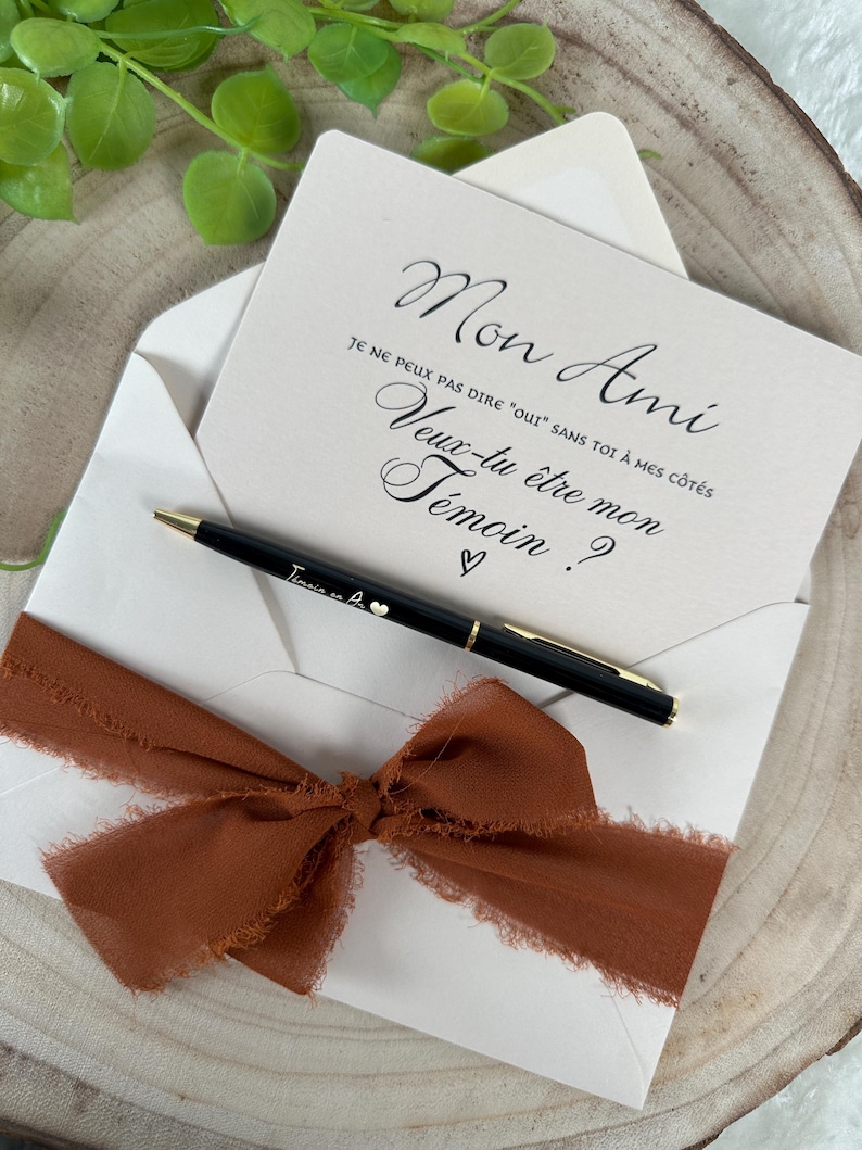 May include: An invitation card with the text "Mon Ami" and "Veux-tu &ecirc;tre mon T&eacute;moin?" in elegant script, inside a white envelope. A black pen with gold accents and a rust-colored ribbon are included.