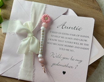 May include: A pink card with the text "Auntie I need an extra pair of hands, to help me learn and grow. I know that yours will be the best because Mummy and Daddy told me so... Will you be my godmother?"  A pink and white beaded bracelet with a pink flower charm is attached to the card.