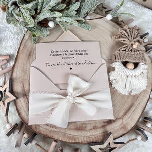 May include: A beige envelope tied with a cream ribbon, containing a card with French text. The card is on a wooden slice, surrounded by decorative elements including a small gnome, wooden stars, and faux greenery.