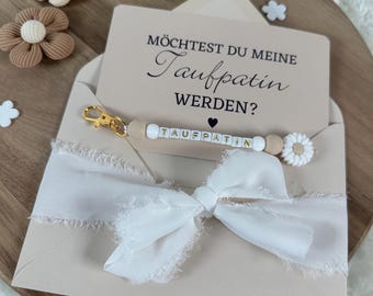 May include: A light brown envelope with a white bow and a card that says "Möchtest du meine Taufpatin werden?"  The card is attached to a white beaded bracelet with the word "TAUFPATIN" spelled out in white letters.