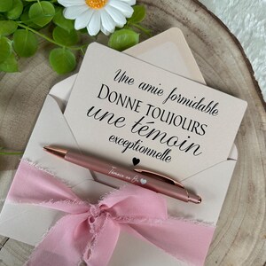 K&ouml;nnte beinhalten: Ein ros&eacute;goldener Stift liegt auf einem cremefarbenen Umschlag, der mit einem rosa Band gebunden ist. Eine Karte im Umschlag hat franz&ouml;sischen Text. Eine wei&szlig;e G&auml;nsebl&uuml;mchen mit gelbem Zentrum und gr&uuml;nen Bl&auml;ttern ist im Hintergrund.