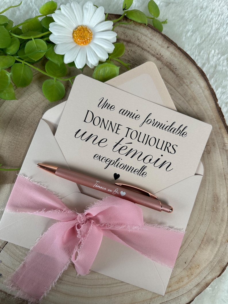 K&ouml;nnte beinhalten: Ein ros&eacute;goldener Stift liegt auf einem cremefarbenen Umschlag, der mit einem rosa Band gebunden ist. Eine Karte im Umschlag hat franz&ouml;sischen Text. Eine wei&szlig;e G&auml;nsebl&uuml;mchen mit gelbem Zentrum und gr&uuml;nen Bl&auml;ttern ist im Hintergrund.