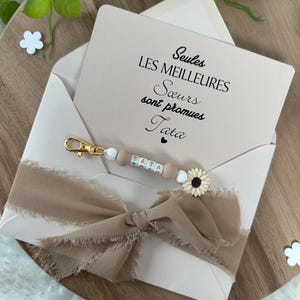 May include: A light pink envelope with a white and brown ribbon tied in a bow. The envelope has a card inside with the text "Seules les meilleures soeurs sont promues Tata".