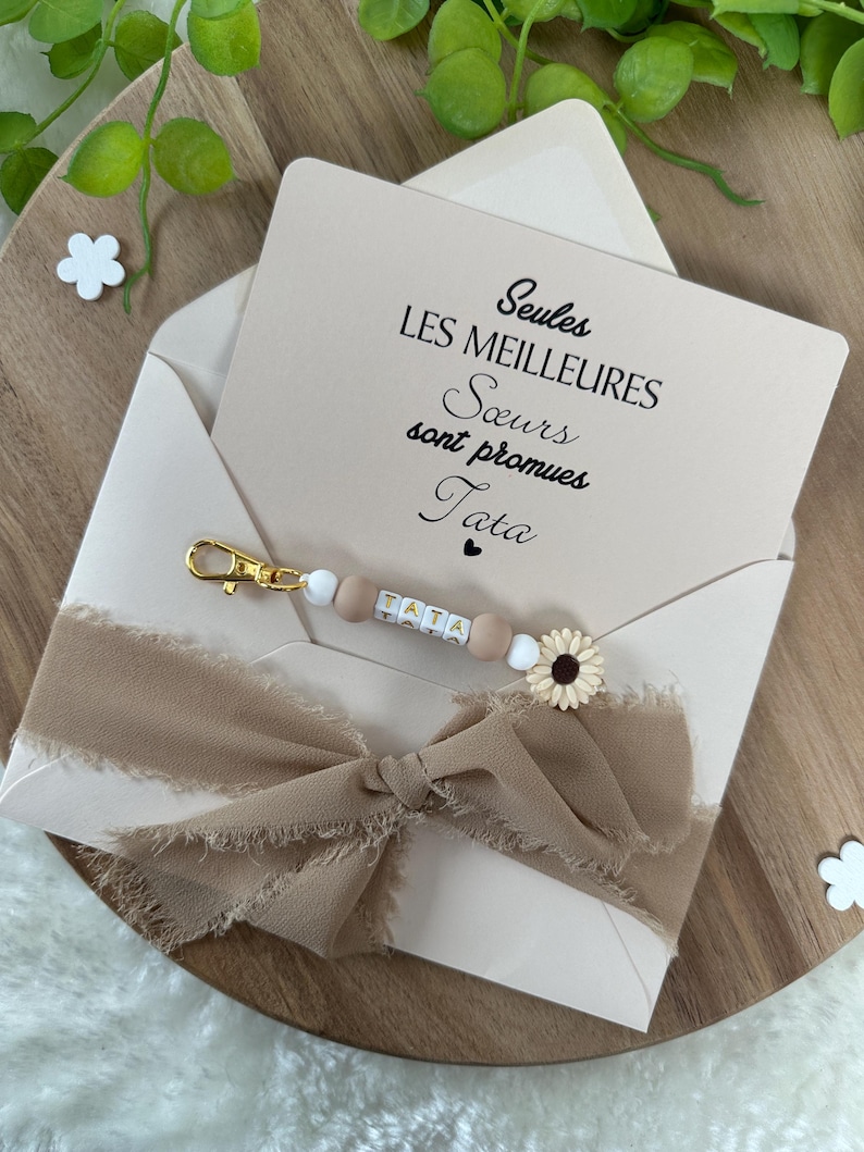 May include: A light pink envelope with a white and brown ribbon tied in a bow. The envelope has a card inside with the text "Seules les meilleures soeurs sont promues Tata".