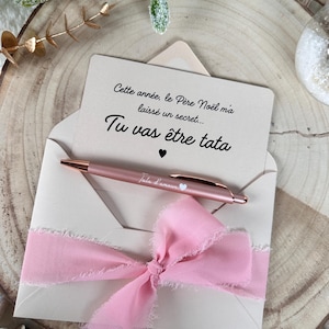 Puede incluir: Un sobre beige claro con una cinta rosa y una tarjeta que dice "Tu vas être tata". Un bolígrafo de oro rosa con el texto "Tata d'amour" está colocado en la tarjeta. La escena está ambientada sobre una superficie de madera con adornos navideños.