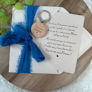 May include: A wooden keychain with the words "Parrain d'amour" and a heart, attached to a silver ring. It is paired with a card and a blue ribbon. The card has French text.