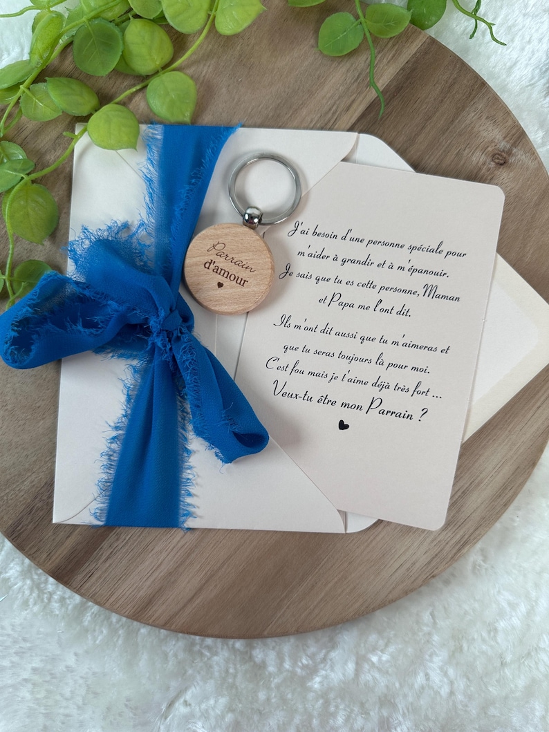 May include: A wooden keychain with the words "Parrain d'amour" and a heart, attached to a silver ring. It is paired with a card and a blue ribbon. The card has French text.