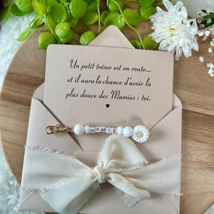 Pu&ograve; includere: Una busta rosa chiaro con un nastro color crema e un portachiavi con la scritta "MAMIE". La carta all'interno dice: "Un petit tr&eacute;sor est en route... et il aura la chance d'avoir la plus douce des Mamies : toi."