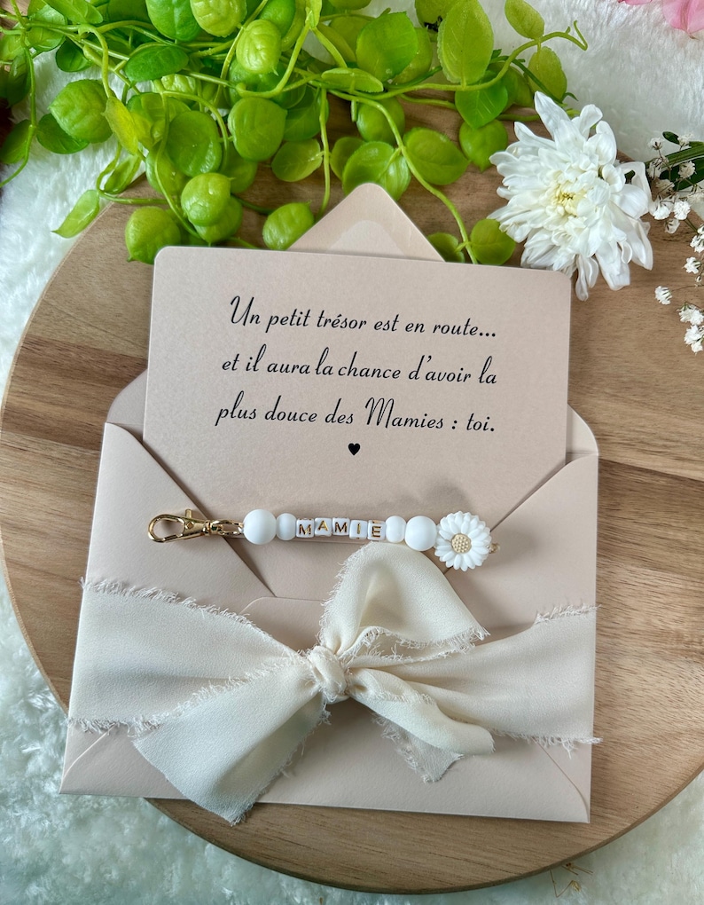 Pu&ograve; includere: Una busta rosa chiaro con un nastro color crema e un portachiavi con la scritta "MAMIE". La carta all'interno dice: "Un petit tr&eacute;sor est en route... et il aura la chance d'avoir la plus douce des Mamies : toi."