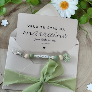 May include: A light brown envelope with a white card inside. The card has the text "VEUX-TU &Ecirc;TRE MA marraine pour toute la vie... (DIS OUI)" in black. A green ribbon is tied around the envelope. A green and white beaded keychain with the word "MARRAINE" spelled out in white letters is attached to the envelope.