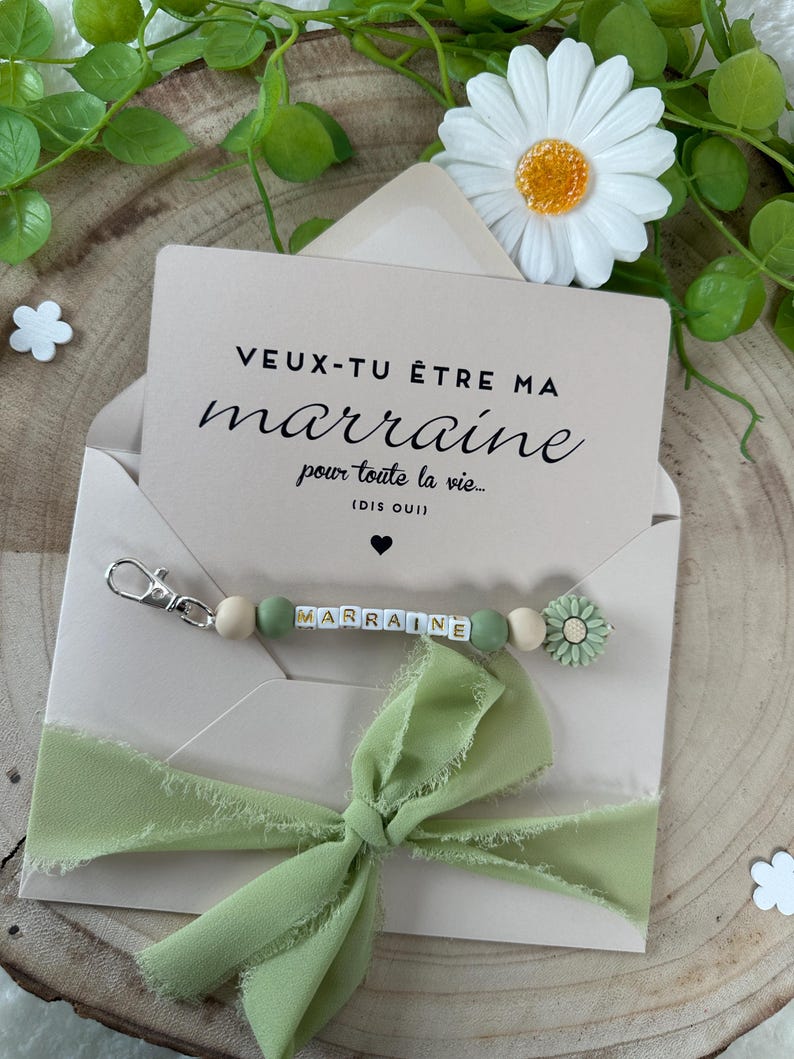 May include: A light brown envelope with a white card inside. The card has the text "VEUX-TU &Ecirc;TRE MA marraine pour toute la vie... (DIS OUI)" in black. A green ribbon is tied around the envelope. A green and white beaded keychain with the word "MARRAINE" spelled out in white letters is attached to the envelope.