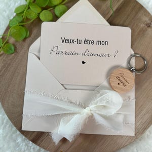 May include: A light beige envelope with a card that reads "Veux-tu &ecirc;tre mon Parrain d'amour?" and a small wooden keychain. The envelope is tied with a white ribbon. The keychain has the same text. The items are on a wooden surface.