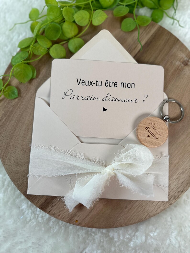 May include: A light beige envelope with a card that reads "Veux-tu &ecirc;tre mon Parrain d'amour?" and a small wooden keychain. The envelope is tied with a white ribbon. The keychain has the same text. The items are on a wooden surface.