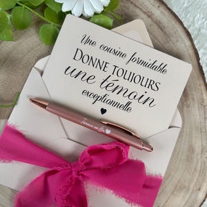 K&ouml;nnte beinhalten: Eine hellrosa Umschlag mit einem pinkfarbenen Band, das eine Gru&szlig;karte mit schwarzer Schrift enth&auml;lt. Ein ros&eacute;goldener Stift mit der Aufschrift "T&eacute;moin en Or" liegt auf dem Umschlag. Eine wei&szlig;e G&auml;nsebl&uuml;mchen und Gr&uuml;nzeug im Hintergrund.