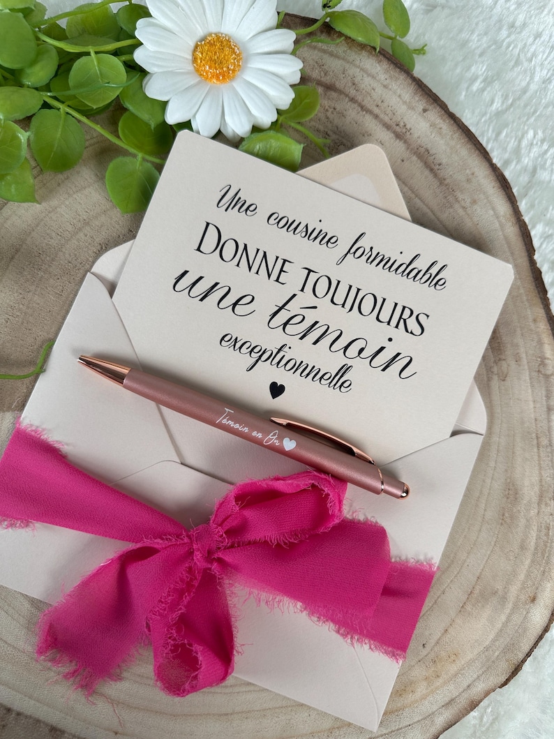 K&ouml;nnte beinhalten: Eine hellrosa Umschlag mit einem pinkfarbenen Band, das eine Gru&szlig;karte mit schwarzer Schrift enth&auml;lt. Ein ros&eacute;goldener Stift mit der Aufschrift "T&eacute;moin en Or" liegt auf dem Umschlag. Eine wei&szlig;e G&auml;nsebl&uuml;mchen und Gr&uuml;nzeug im Hintergrund.