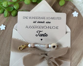 May include: A beige envelope with a card that reads "Eine wunderbare Schwester ist immer eine aussergewöhnliche Tante." A beaded keychain with the word "TANTE" is attached with a gold clasp. The envelope is tied with a beige ribbon.
