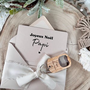 May include: A beige envelope with a white ribbon bow, holding a card that reads "Joyeux Noël Papi". A wooden bottle opener with a keychain is attached. The scene is set on a wooden slice with festive decorations.