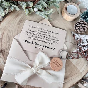 Puede incluir: Un llavero de madera con la inscripción "Marraine d'amour" está sujeto a un sobre beige con una cinta blanca. Dentro hay una tarjeta con texto en francés. La escena está ambientada sobre una rodaja de madera con adornos navideños.
