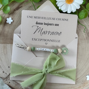 May include: A light green envelope with a white card inside. The card says "UNE MERVEILLEUSE COUSINE donne toujours une Marraine EXCEPTIONNELLE". A light green ribbon is tied around the envelope. A beaded keychain with the word "MARRAINE" spelled out in white letters on green beads is on top of the envelope.