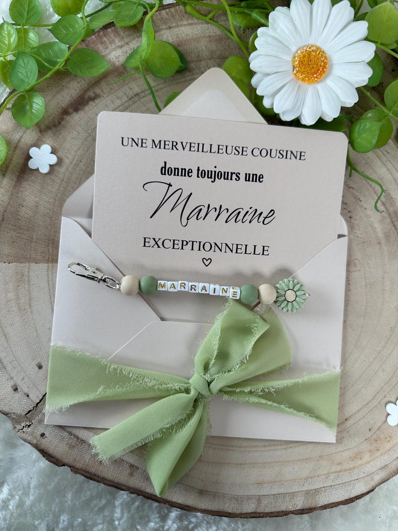 May include: A light green envelope with a white card inside. The card says "UNE MERVEILLEUSE COUSINE donne toujours une Marraine EXCEPTIONNELLE". A light green ribbon is tied around the envelope. A beaded keychain with the word "MARRAINE" spelled out in white letters on green beads is on top of the envelope.