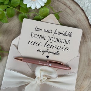 K&ouml;nnte beinhalten: Ein ros&eacute;goldener Stift und eine Karte mit franz&ouml;sischem Text werden in einem hellrosa Umschlag mit cremefarbenem Band pr&auml;sentiert. Auf der Karte steht: "Une s&oelig;ur formidable donne toujours une t&eacute;moin exceptionnelle."
